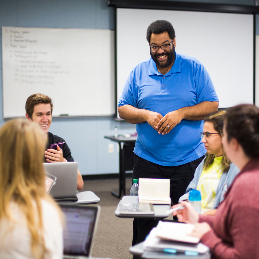 Professor in blue shirt smiles at group of students sitting in a circle